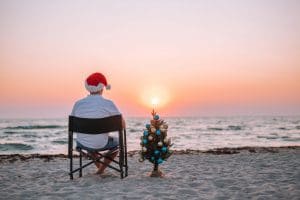A guy in a santa hat sits on the beach, on a chair overlooking the sea and enjoys the sunset, next to a tree decorated with Christmas balls.